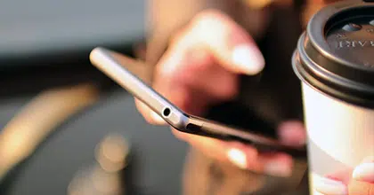 Eine Hand hält Handy, in der anderen sieht man einen Coffee To Go Becher